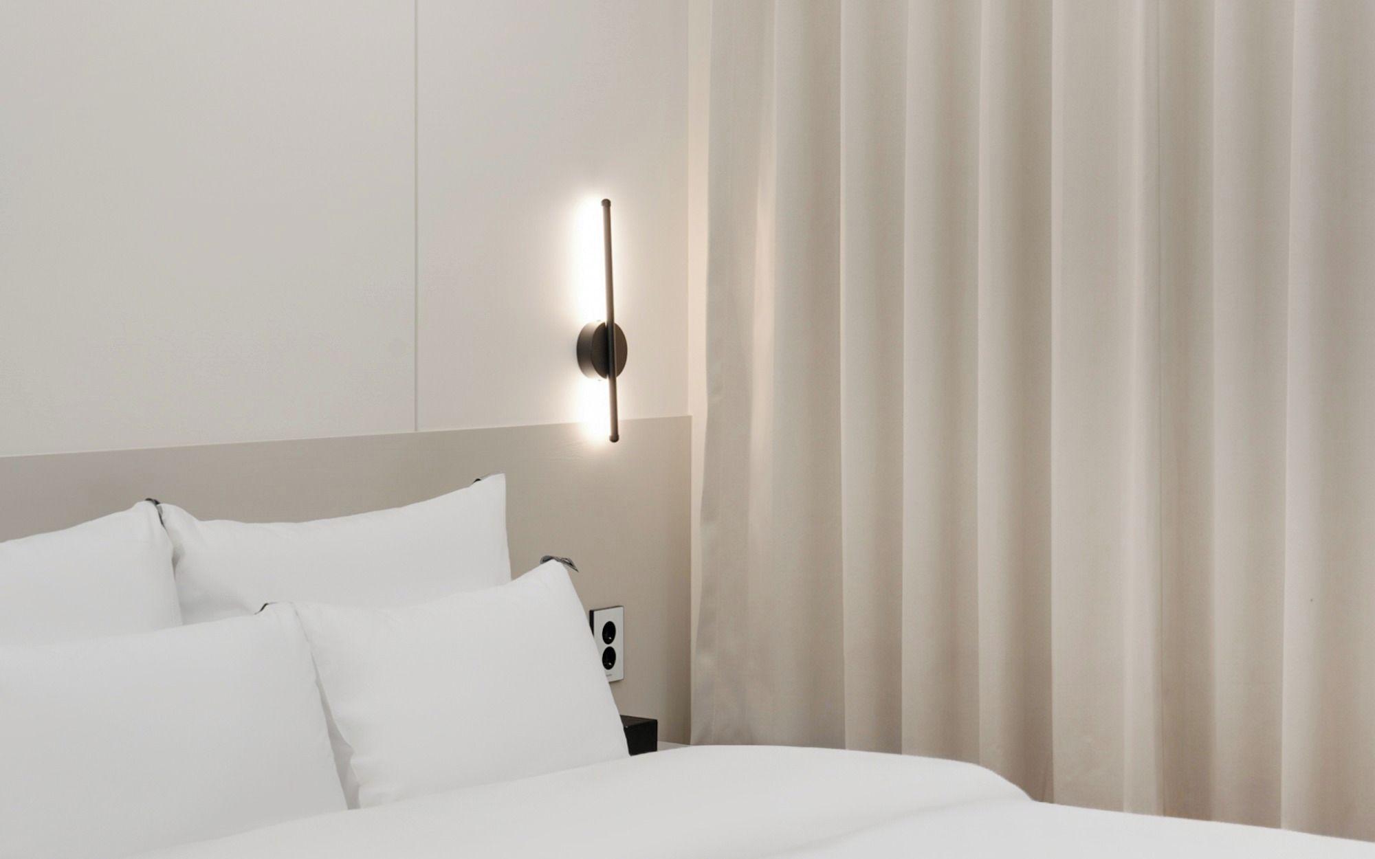 A hotel room with a bed, white linens, a wall-mounted light fixture, and a curtain.