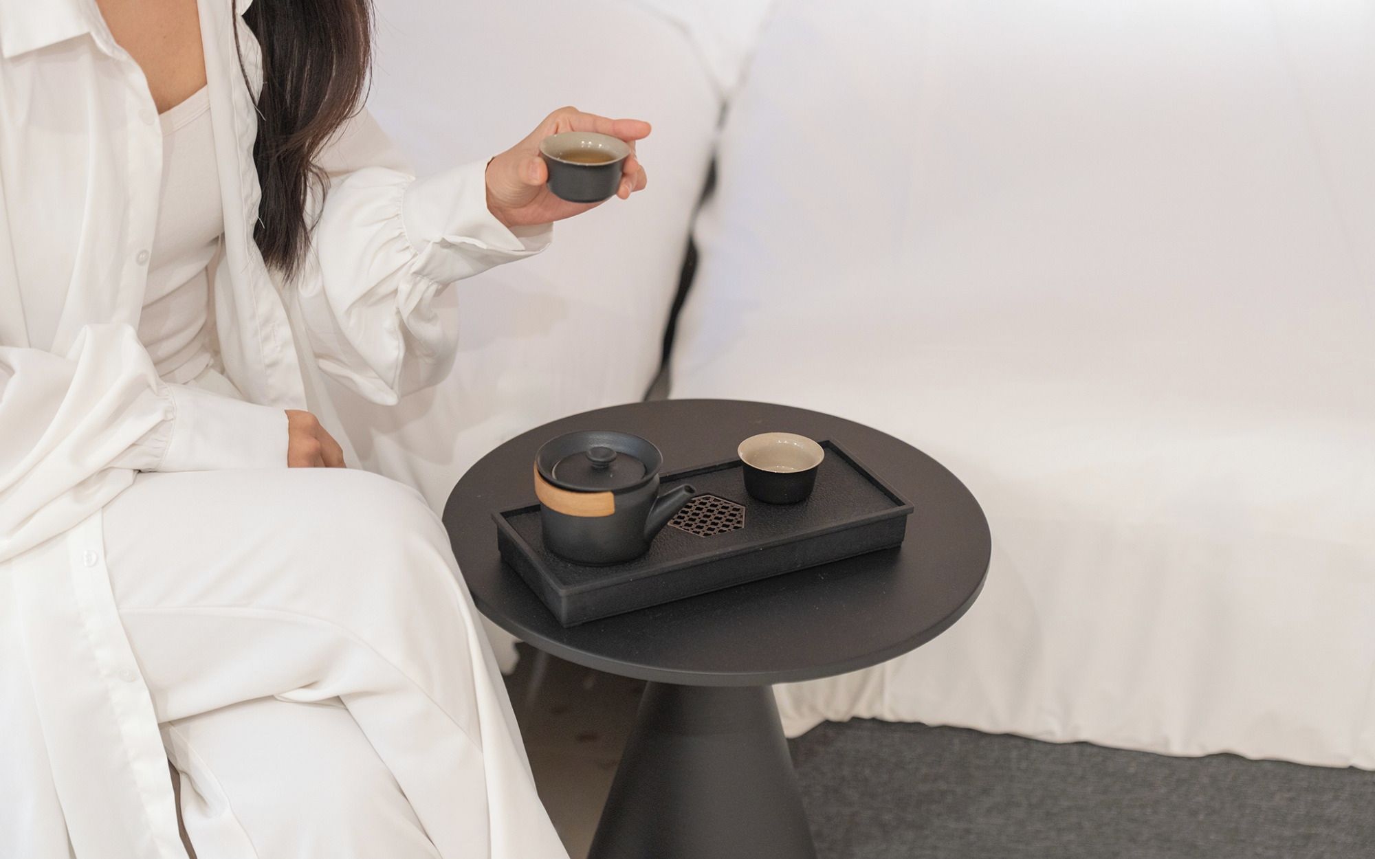 Eine Person sitzt auf einem weißen Sofa mit einem schwarzen Tablett, das einen Teekessel, eine Tasse und eine kleine Schüssel enthält.