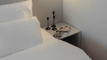 Een keurig opgemaakt bed met witte lakens, een nachtkastje met een boek, een kopje en twee zwarte kandelaars.