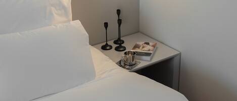 A neatly made bed with white linens, a bedside table with a book, a cup, and two black candlesticks.