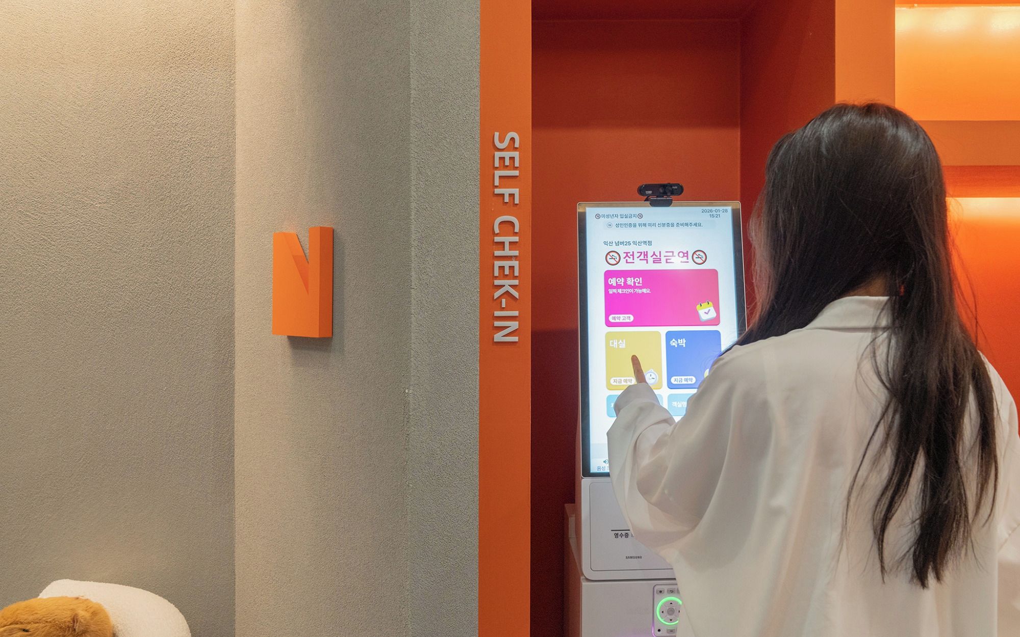 A person interacting with a self-service kiosk in a room with a beige wall and an orange accent wall.