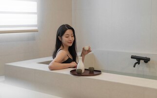 Una persona sentada en una bañera, con una bandeja que contiene un jarrón blanco y dos tazas pequeñas.