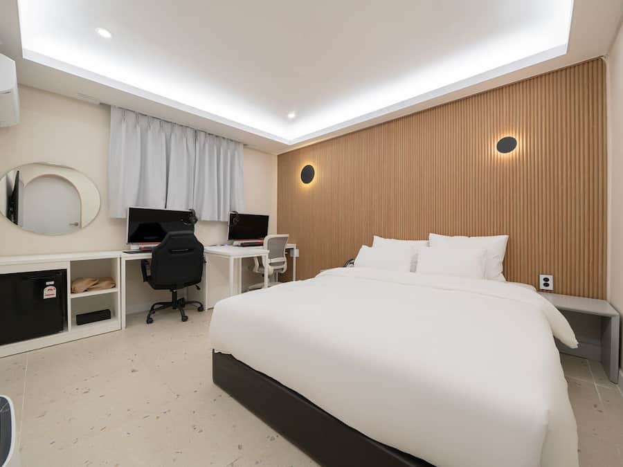 Quarto de hotel moderno com uma cama grande, uma escrivaninha com dois monitores, uma cadeira e um guarda-roupa.