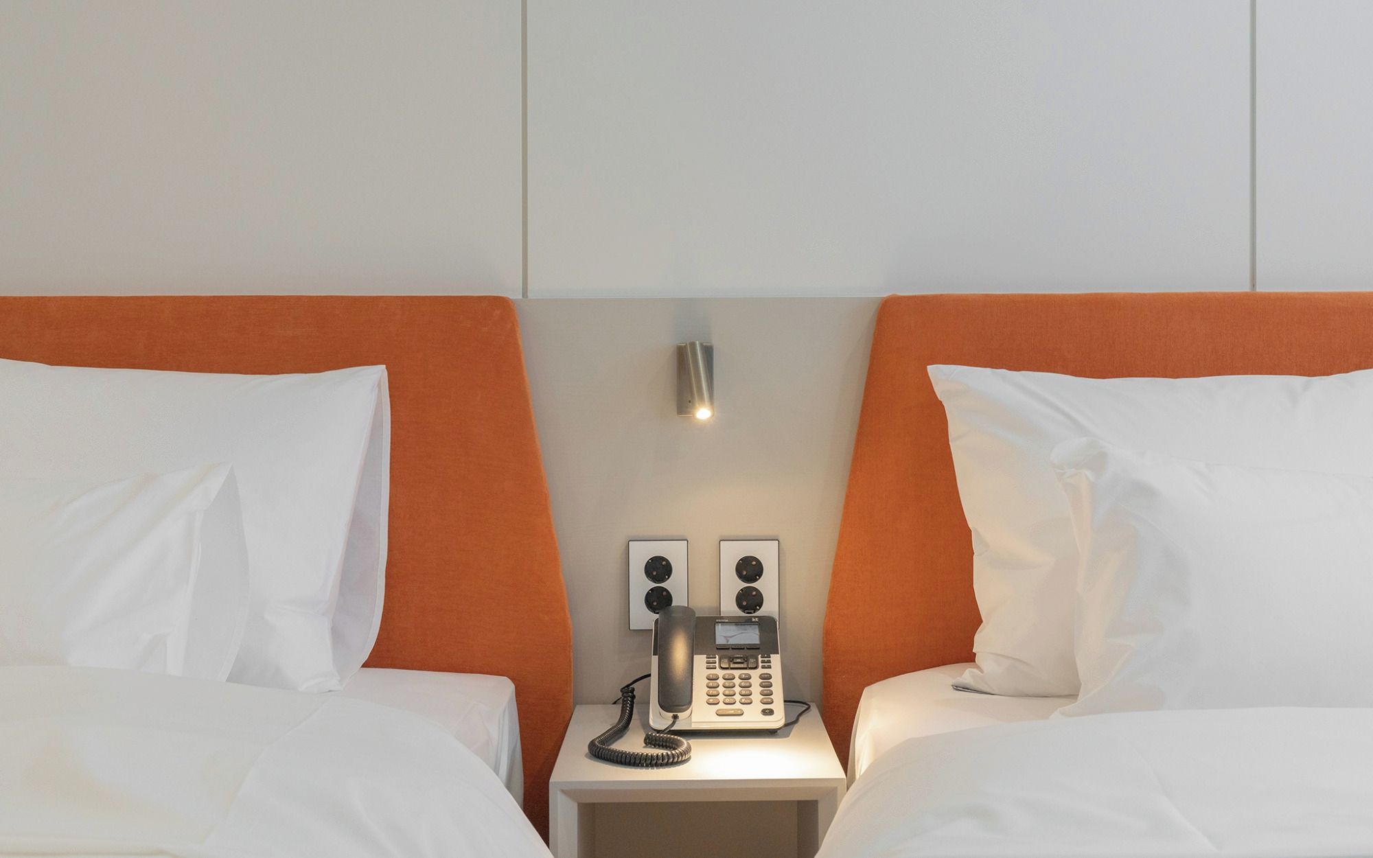 A hotel room with two beds, a nightstand with a telephone, and a wall-mounted light fixture.