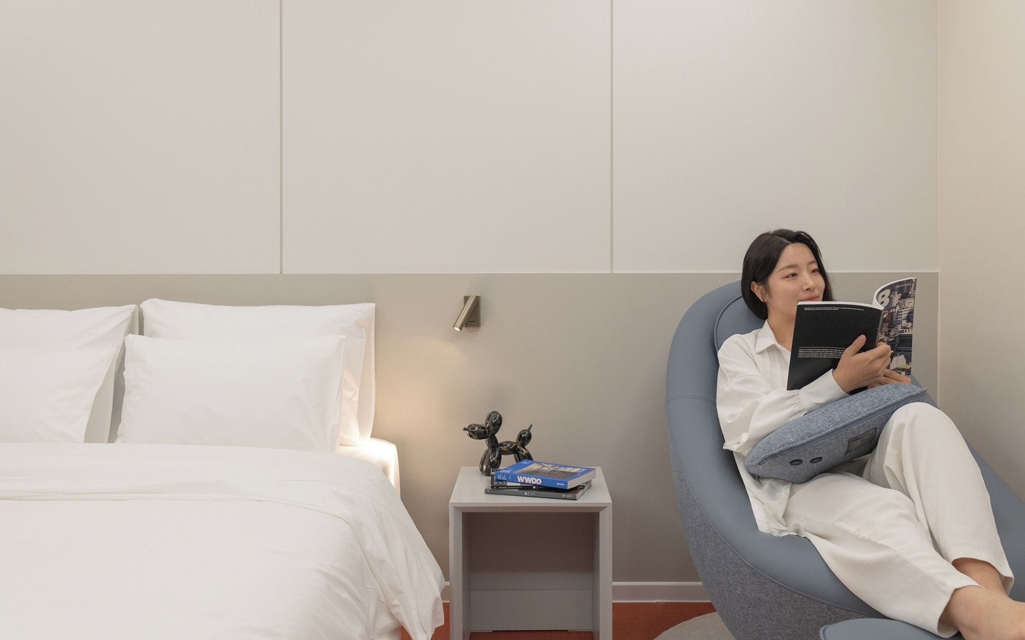 Una persona sentada en una silla leyendo un libro en una habitación con una cama y una pequeña mesa llena de libros.