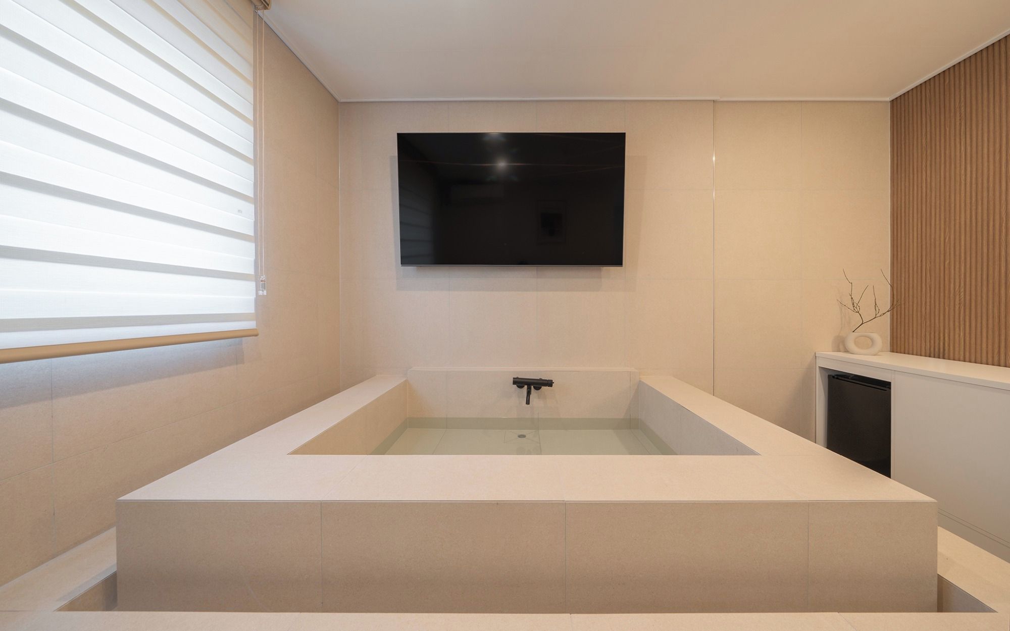 A modern bathroom with a freestanding bathtub, a wall-mounted TV, and a window with blinds.