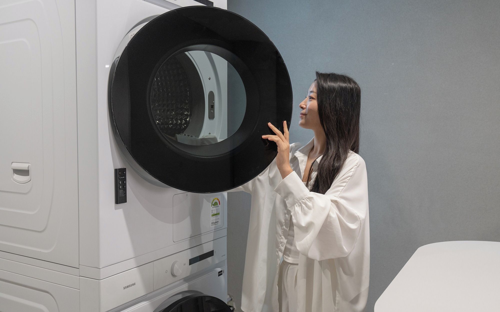 A person holding a circular object in front of a white appliance, possibly a washing machine, with a control panel and a brand label visible.