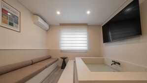A modern bathroom with a large bathtub, a bench, a small table, and a window with blinds.