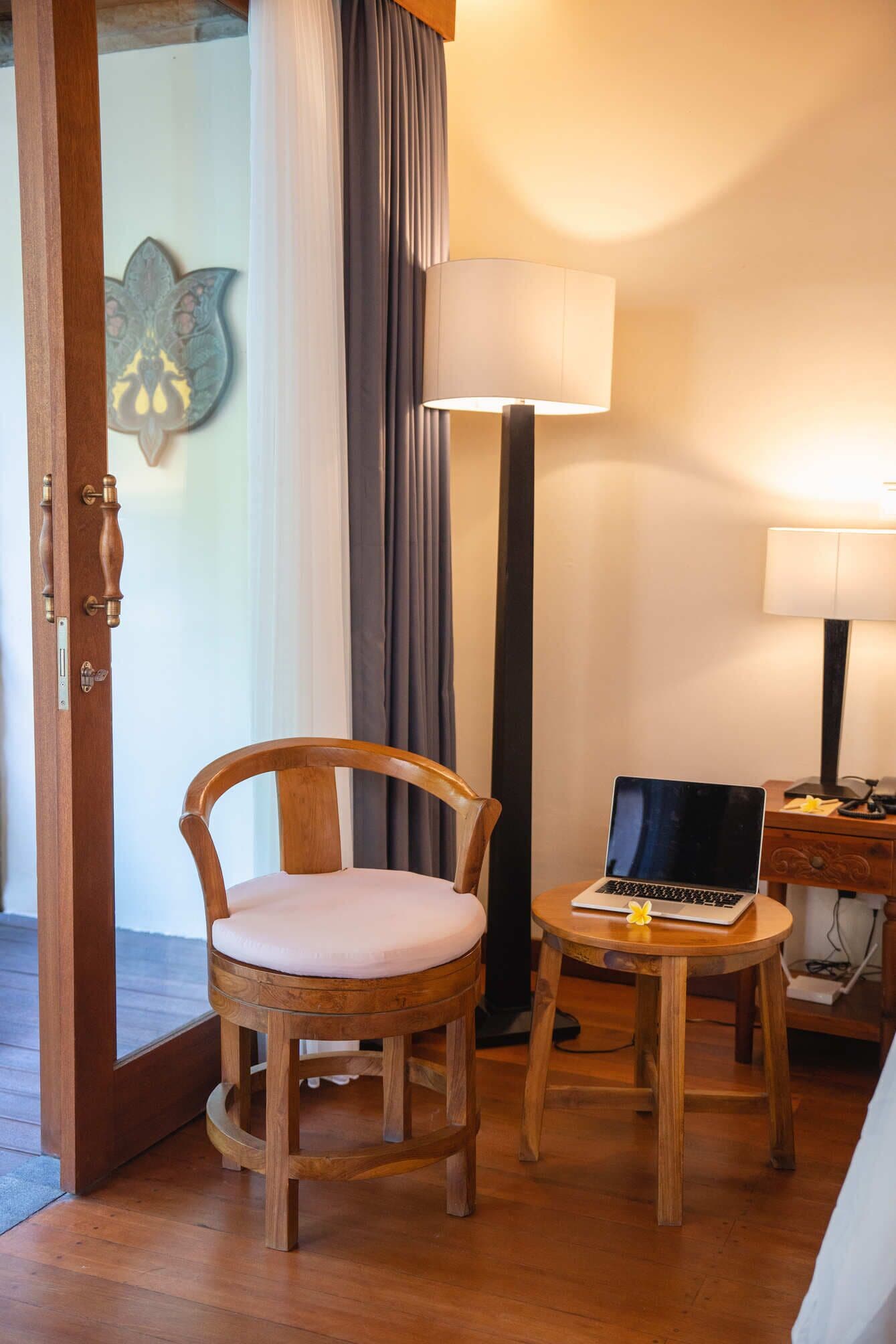 Une chaise en bois avec un coussin, un ordinateur portable posé sur une petite table, une lampe et une porte ornée d’une plaque décorative.