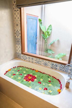A bathroom with a bathtub filled with water and floating flowers, a window with a view of a door and plants outside, and a tiled wall with a decorative border. - The Compass Rose Ubud (Ubud)