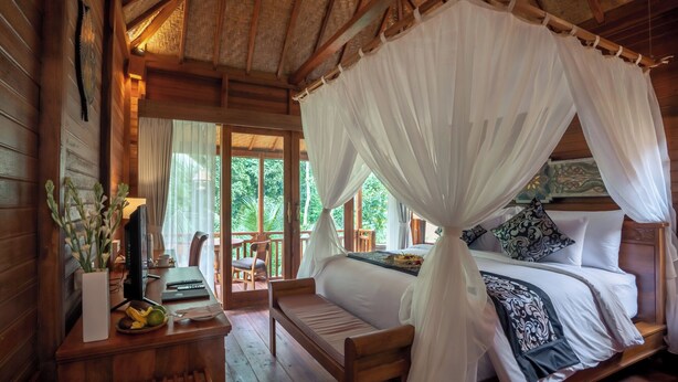 Una habitación de madera con una cama bajo un dosel, un escritorio con un ordenador y vistas a la vegetación exterior.