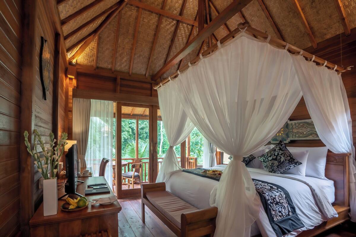 A wooden room with a bed under a canopy, a desk with a computer, and a view of greenery outside.