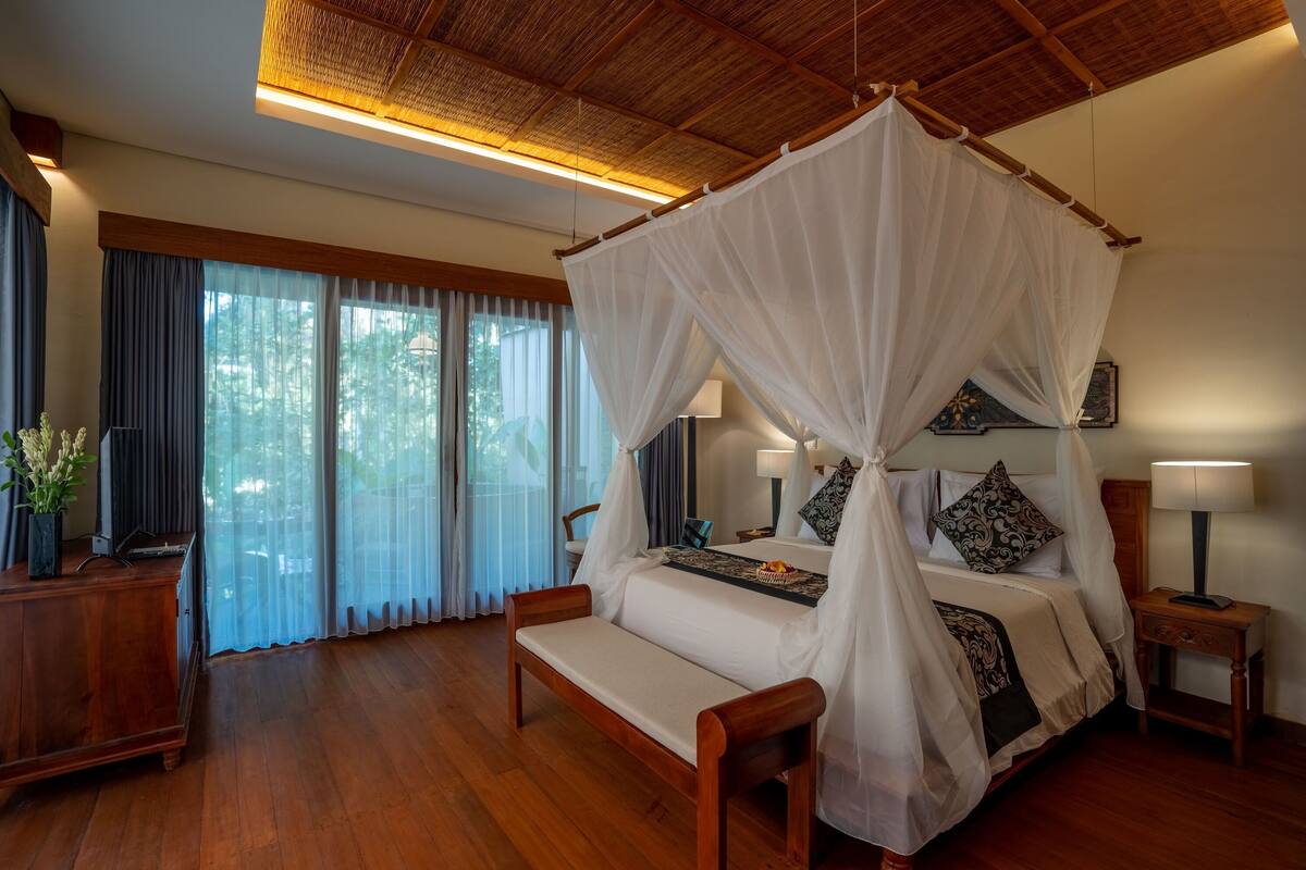A four-poster bed with a canopy, bedside lamps, a wooden bench, and a view of the outdoors through large windows.