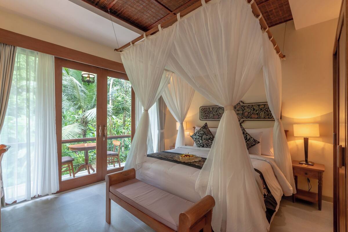 A bedroom with a canopy bed, a bench, a nightstand, and a view of lush greenery outside.