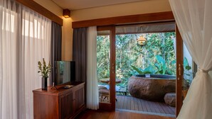 Una habitación con un armario de madera, un televisor, una zona de comedor con mesa y sillas, y vistas a un jardín con rocas y plantas.