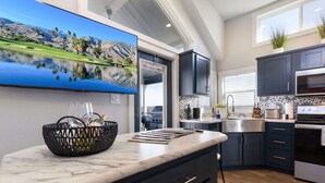 A modern kitchen with dark cabinets, a marble countertop, and a large TV displaying a scenic landscape. - Bluemoon Retreat - Tiny Home 115 (Fredericksburg)