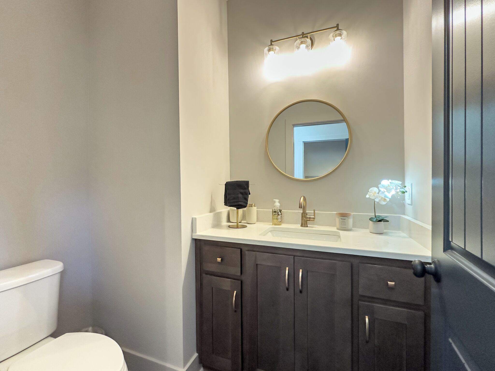 A bathroom with a white toilet, dark wooden cabinets, a round mirror, and a white sink.