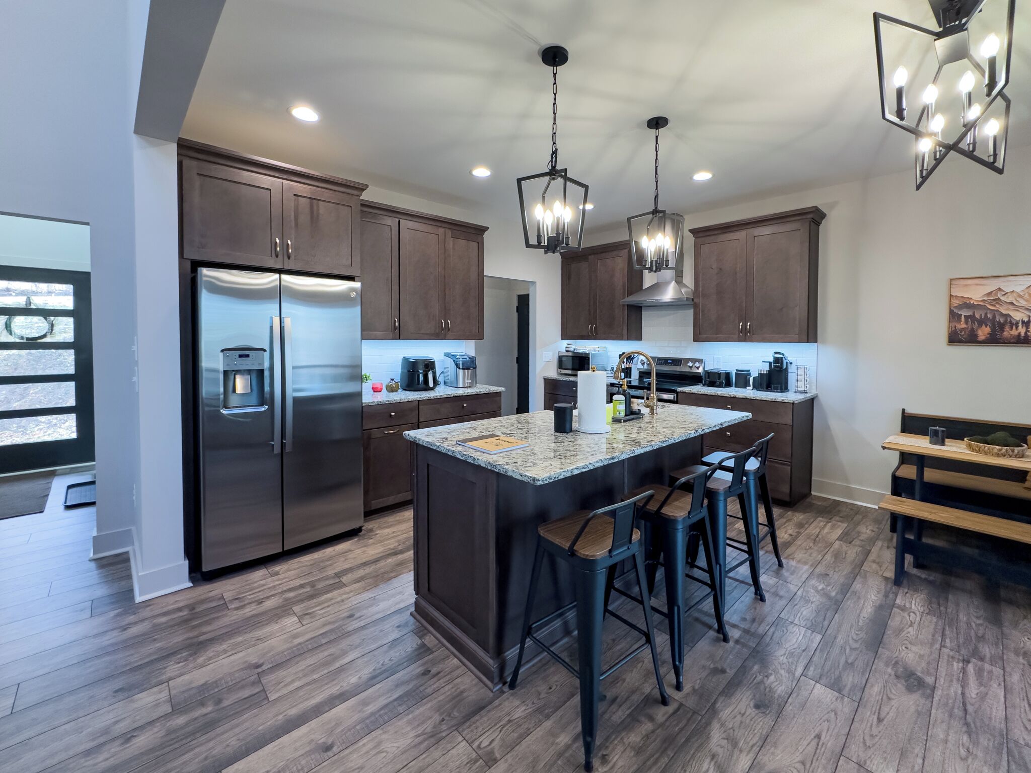 A modern kitchen with dark wood cabinets, a stainless steel refrigerator, a granite countertop island with bar stools, and pendant lighting.