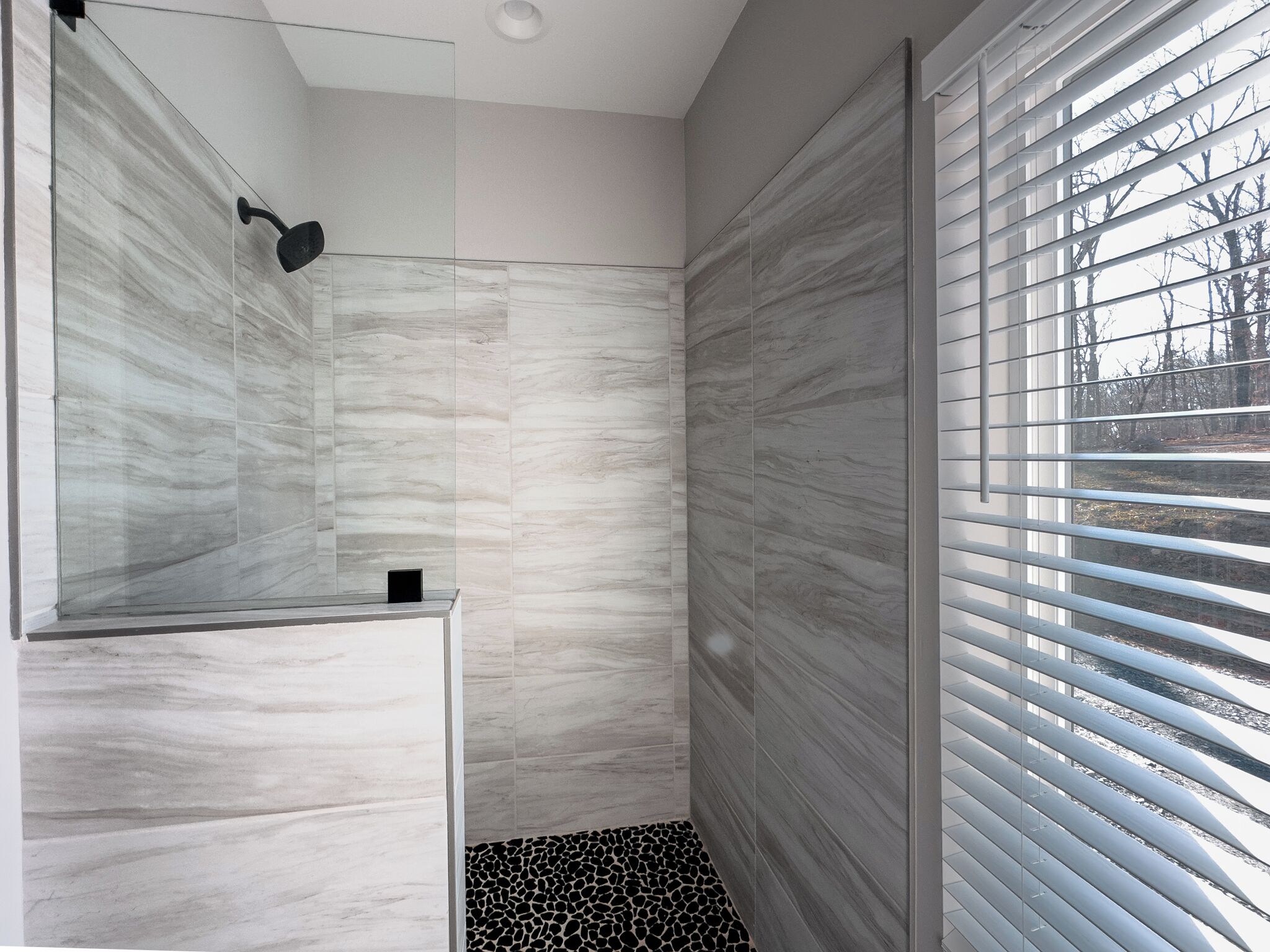 A modern bathroom with a glass shower enclosure, a marble wall, and a view through window blinds.