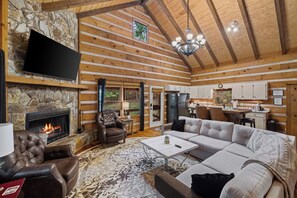 Ein Blockhaus-Interieur mit Kamin aus Stein, Ledersesseln, einem großen Sofa, einem Fernseher und einer Küchenzeile.