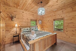 Ein Schlafzimmer einer Holzhütte mit einem Bett, einem Nachttisch, einer Lampe, einer Dekoration mit einem Hirschkopf und einem Fenster mit Blick auf Bäume.