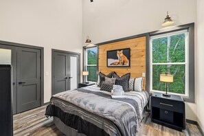 Ein Schlafzimmer mit einem Bett, einem Nachttisch, einem Wandbild und einem Fenster mit Blick auf Bäume.