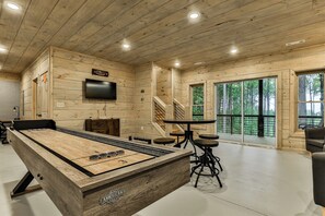 A wooden interior with a pool table, a TV, and a staircase leading to a balcony with a forest view. - Knotty by Nature | Cherry Log Mountain Cabin (Blue Ridge)