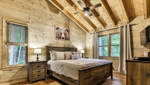 A wooden cabin bedroom with a bed, nightstands, a ceiling fan, and large windows. - Knotty by Nature | Cherry Log Mountain Cabin (Blue Ridge)