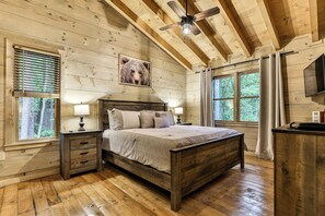 A wooden cabin bedroom with a bed, nightstands, a ceiling fan, and large windows. - Knotty by Nature | Cherry Log Mountain Cabin (Blue Ridge)