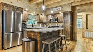 A rustic kitchen with wooden cabinets, stainless steel appliances, and a bar area with stools. - Knotty by Nature | Cherry Log Mountain Cabin (Blue Ridge)