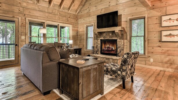 A log cabin interior with a stone fireplace, a sofa, a patterned armchair, and framed fish artwork on the walls. - Knotty by Nature | Cherry Log Mountain Cabin (Blue Ridge)