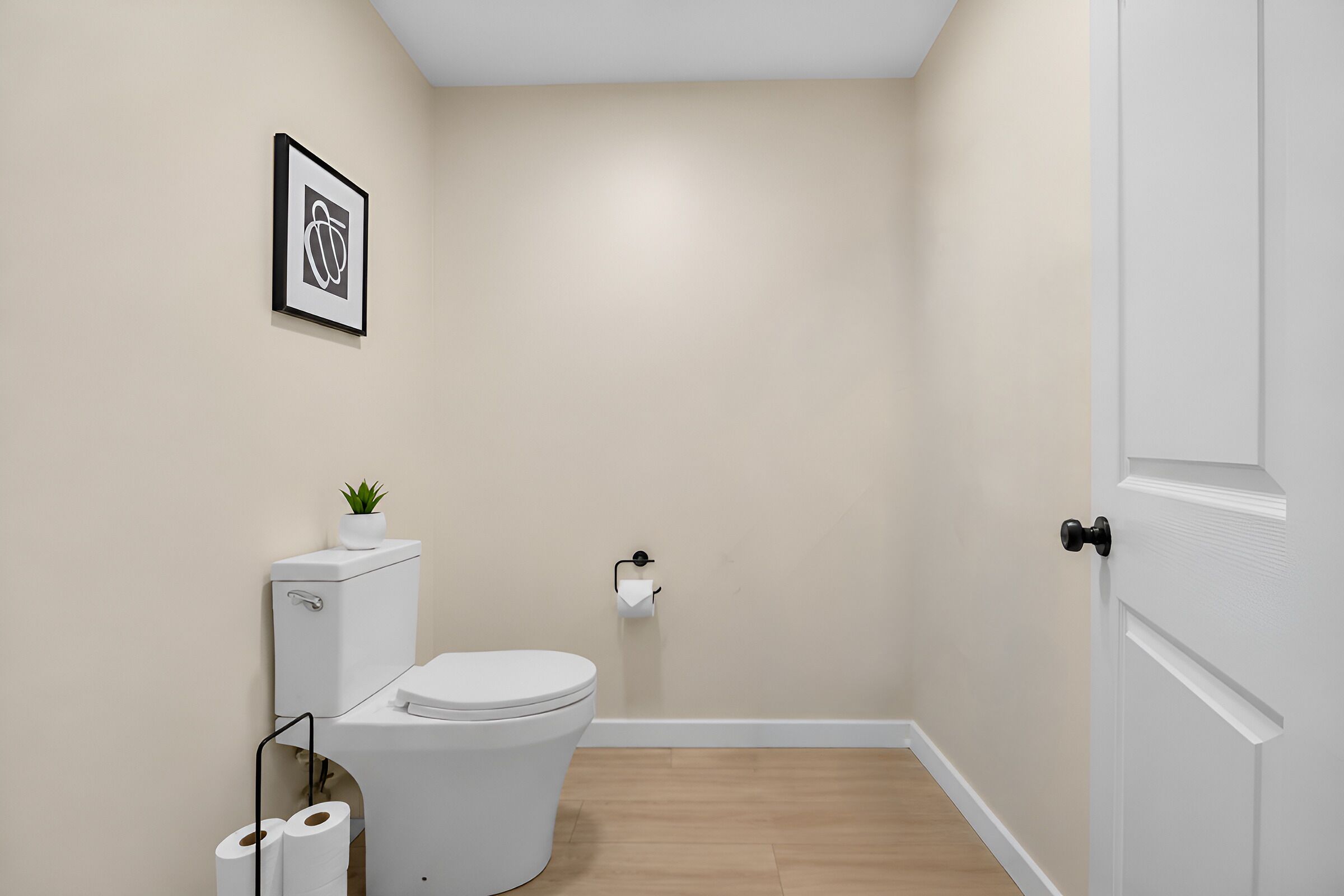 Un baño pequeño y moderno con un inodoro, un rollo de papel higiénico, un cuadro en la pared y una puerta.