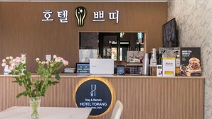 Ein Hotelrezeptionstresen im modernen Design, mit einem Logo, einem Schild mit koreanischen Schriftzeichen und einem Schild für 'Stay & Ramen HOTEL TORANG'. Auf der Theke stehen eine Vase mit Blumen, ein Samsung-Gerät und eine Maxim-Kaffeemaschine.