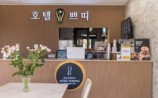 Un mostrador de recepción de hotel con diseño moderno, que incluye un logotipo, un cartel con caracteres coreanos y un cartel que dice 'Stay & Ramen HOTEL TORANG'. Hay una jarra con flores, un dispositivo Samsung y una cafetera Maxim sobre el mostrador.
