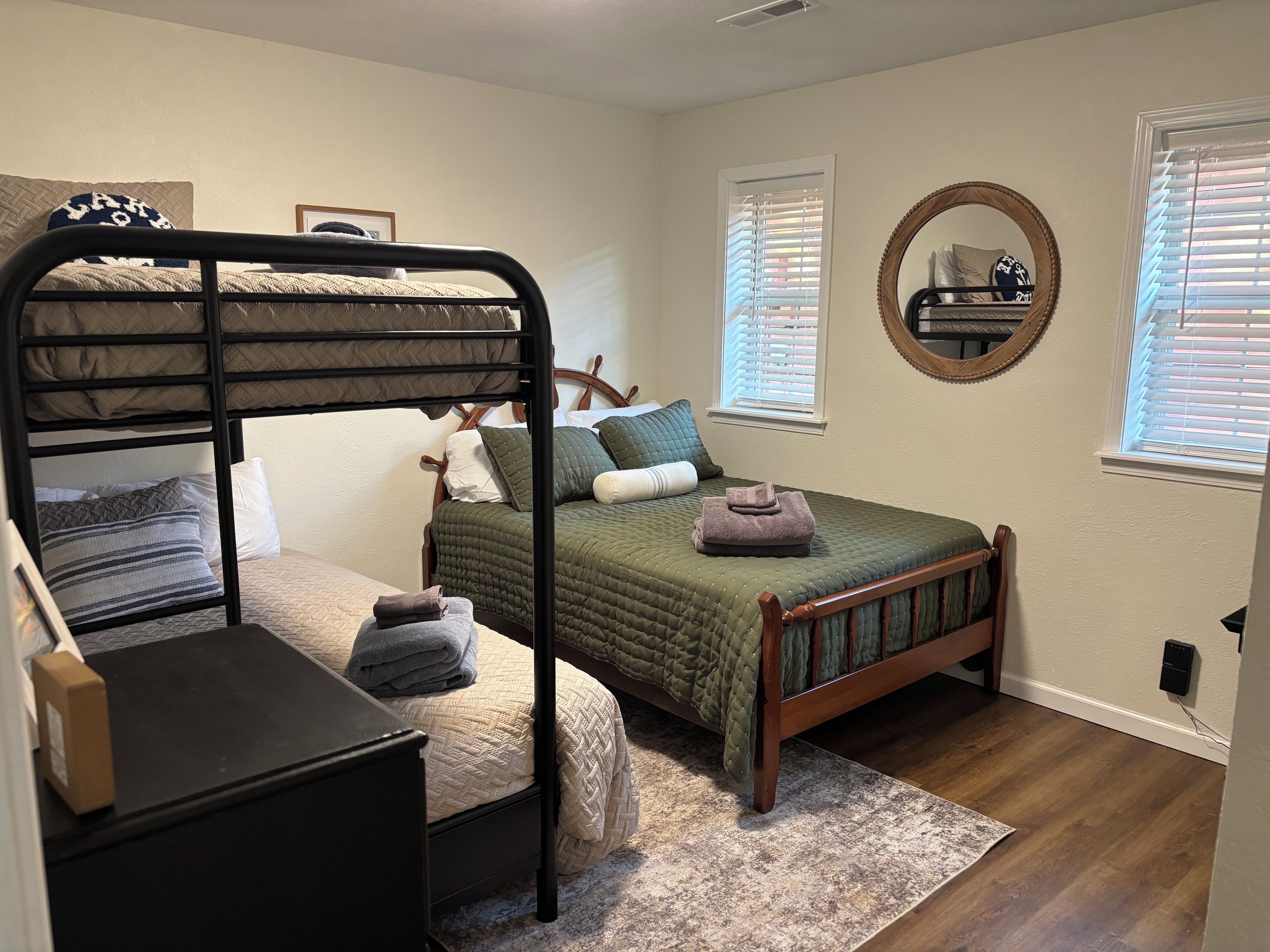 A bedroom with bunk beds, a bedside table, a chair, a window with blinds, a mirror, and a wall-mounted shelf.