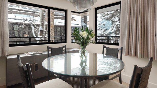 A round glass table with chairs, a vase with flowers, and a view of snow-covered trees through large windows. - Ski-In Aspen Retreat Next To Lift 1A with Fireplace & Walkable Downtown Access (Aspen)