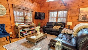A cozy cabin interior with a leather sofa, a coffee table, a bookshelf, and a television mounted on the wall.