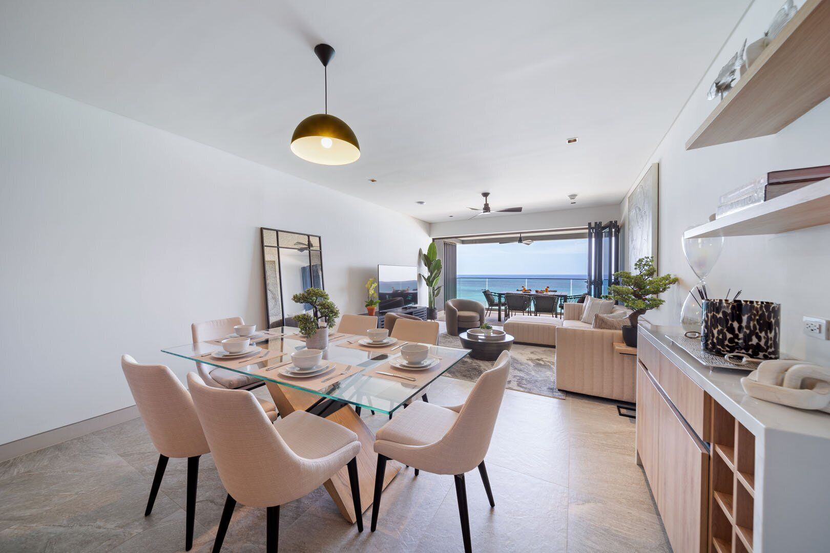 A modern living room with a glass dining table, beige chairs, and a view of the ocean.