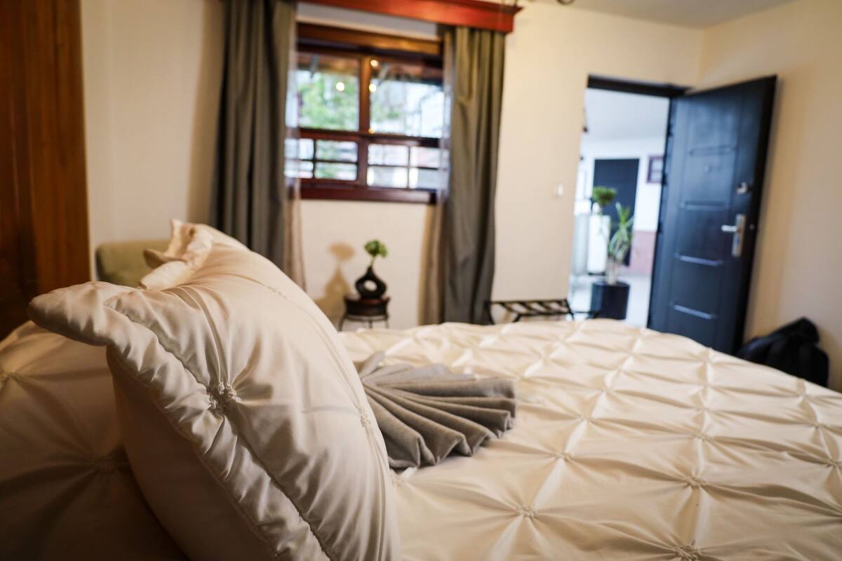 Ein ordentlich bezogenes Bett mit einem gesteppten Kopfteil und Kissen, ein Fenster mit Vorhängen und eine Tür im Hintergrund.
