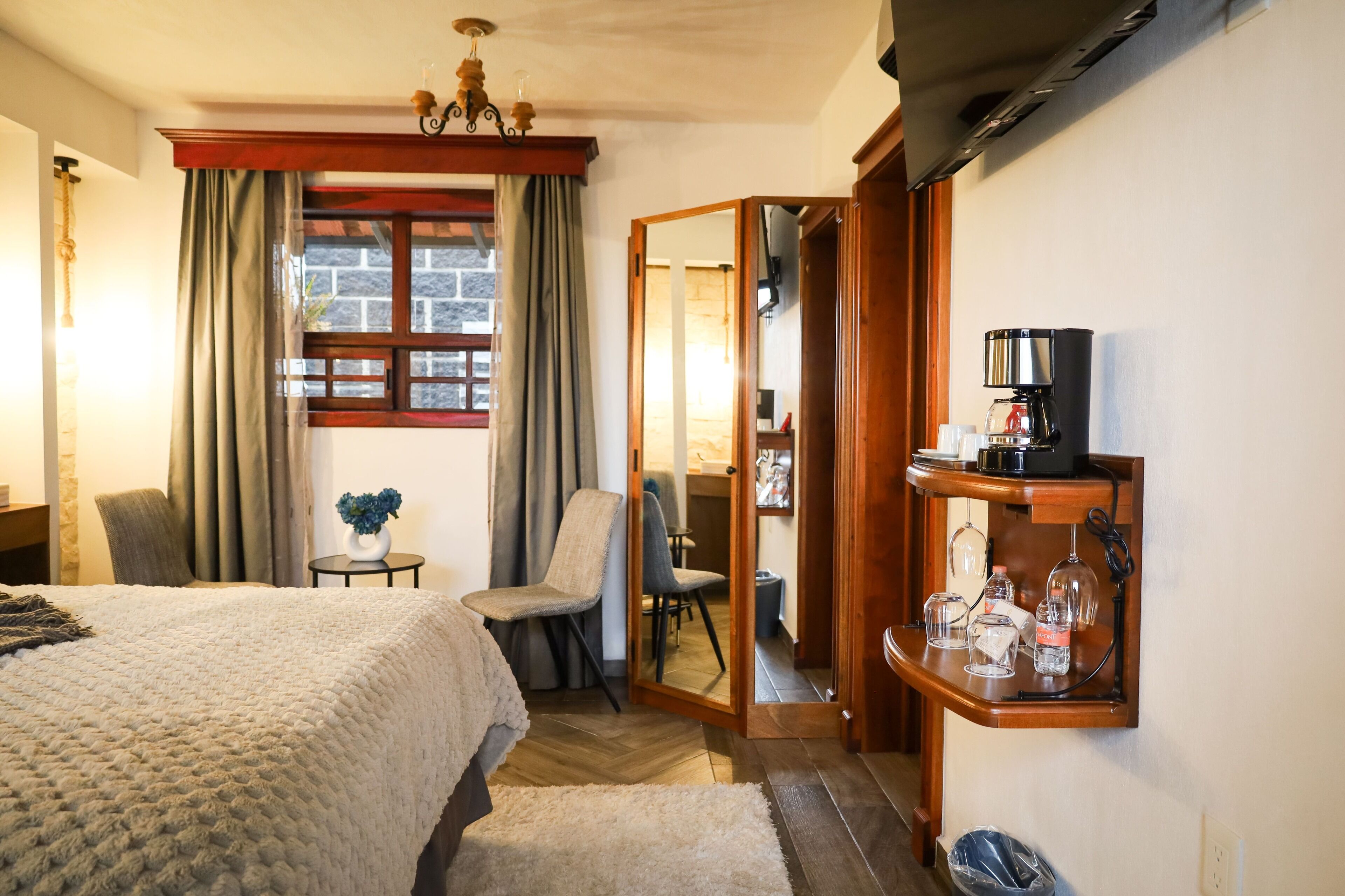 Habitación de hotel con cama, una silla, una mesita con un florero, una cafetera y un espejo.