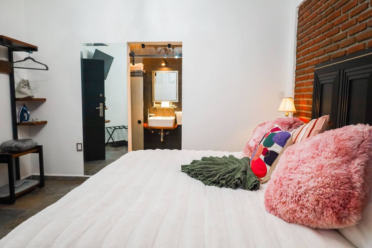 Ein Schlafzimmer mit Bett, Hängesessel, einer Regalkonstruktion mit Gegenständen und einem durchgehenden Türrahmen sichtbarem Badezimmer.