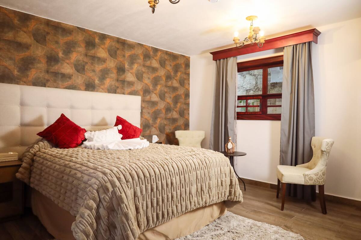 Ein Schlafzimmer mit einem großen Bett, einem Nachttisch, einem Sessel und einem Fenster mit Vorhängen.