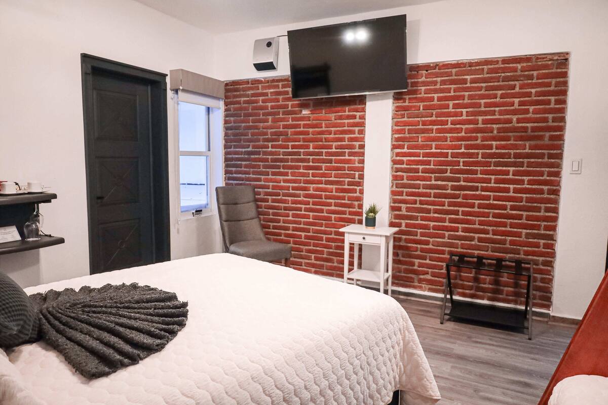 Ein Schlafzimmer mit Backsteinwand, einem flachbildfernseher über dem Bett, einem Sessel, einem kleinen Tisch mit Pflanze und einem Fenster mit Aussicht.