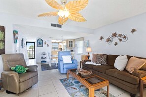 A living room with a ceiling fan, a brown sofa, a blue armchair, a wooden coffee table, and a kitchen in the background. - Charming Cocoa Beach Condo w/ Private Balcony, Heated Pool, Putting Green, & BBQ Grill! ☀️🌴 (Cocoa Beach)