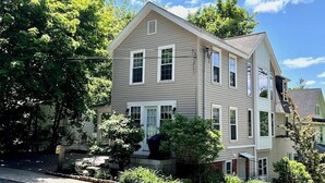 A two-story house with a front porch, a garage, and a well-maintained garden with flowering plants. - Pristine 3 Bedroom, Walk-to Gt Barrington Center! (Great Barrington)