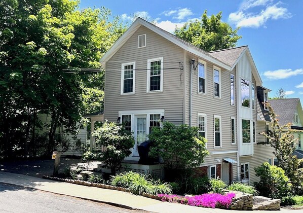 A two-story house with a front porch, a garage, and a well-maintained garden with flowering plants. - Pristine 3 Bedroom, Walk-to Gt Barrington Center! (Great Barrington)