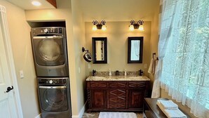 A compact laundry room with two stacked washers and dryers, a vanity with a mirror, and a window with sheer curtains. - Pristine 3 Bedroom, Walk-to Gt Barrington Center! (Great Barrington)