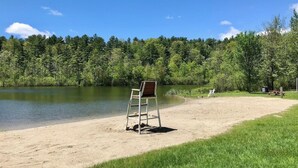 A beach with a sand chair and a picnic table, surrounded by a forest and a lake. - Pristine 3 Bedroom, Walk-to Gt Barrington Center! (Great Barrington)
