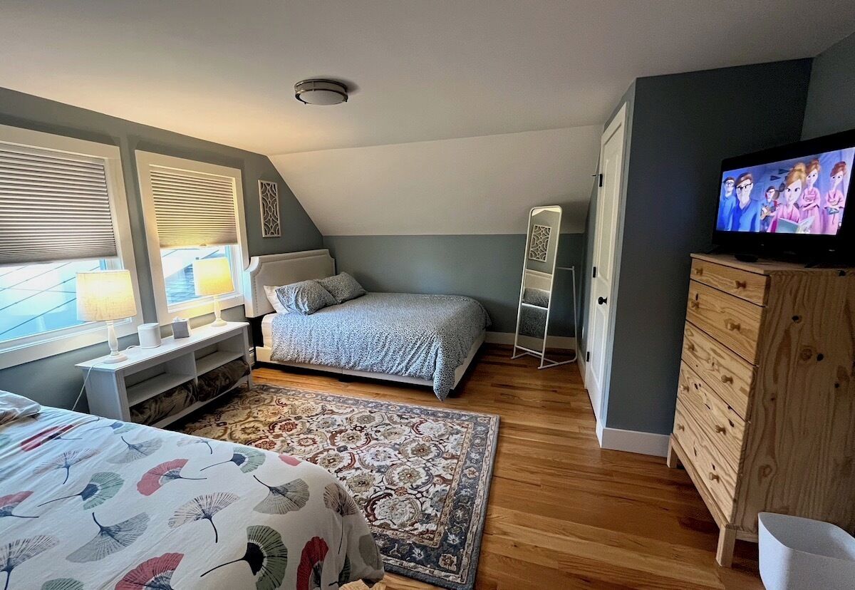 Ein Schlafzimmer mit Bett, Nachttisch, Lampe, Fernseher, Kommode und einem gemusterten Teppich.