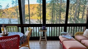A wooden deck with wicker furniture, a small table, and a view of a lake and forest. - Direct on Lake Garfield: Book Fall Weekends! (Monterey)